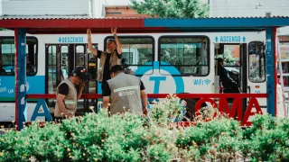 Tres trabajadores con chalecos revisan un colectivo urbano blanco y azul en una parada cubierta al aire libre, con vegetación en primer plano