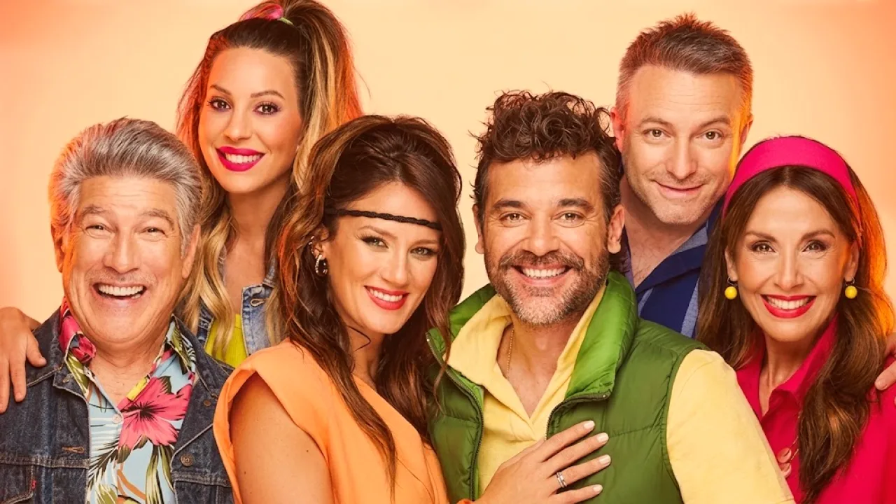 Seis actores sonrientes posan juntos, cinco de ellos con vestimenta colorida, uno con camisa clara y remera debajo, en un fondo neutro.