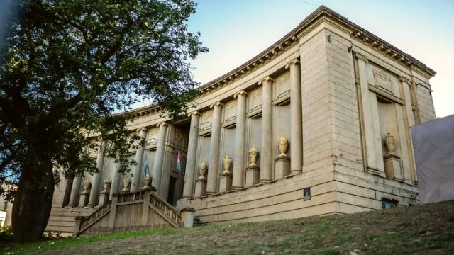 Edificio histórico de estilo clásico con columnas y escalinata principal, ubicado en un entorno con césped y un árbol grande a la izquierda