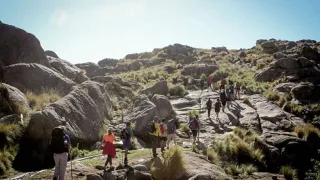 Grupo de personas caminando por sendero rocoso entre montañas y vegetación baja bajo cielo despejado