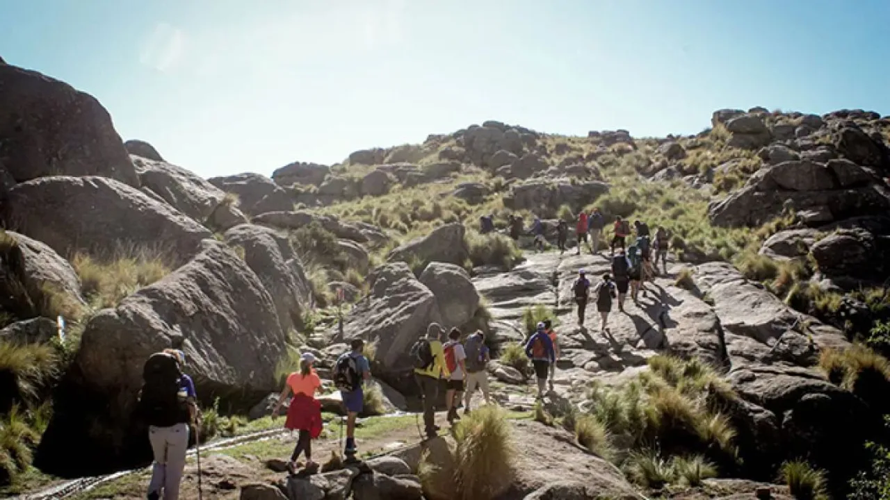 Grupo de personas caminando por sendero rocoso entre montañas y vegetación baja bajo cielo despejado