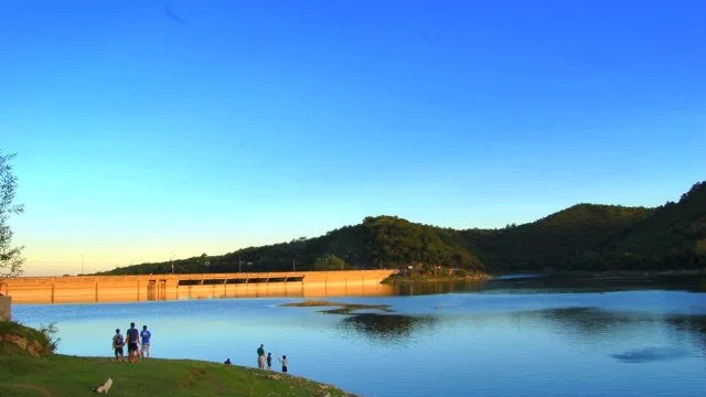 Embalse con agua tranquila, sierra al fondo y personas caminando y paradas sobre un espacio verde con césped
