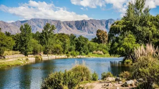 Río con agua tranquila rodeado de árboles y arbustos, con sierras al fondo bajo un cielo parcialmente nublado