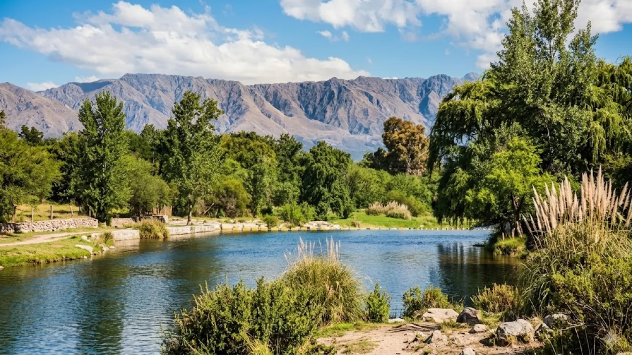 Río con agua tranquila rodeado de árboles y arbustos, con sierras al fondo bajo un cielo parcialmente nublado