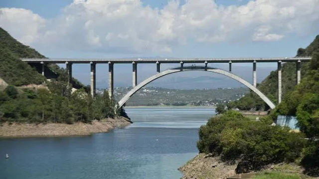 Puente de concreto con un arco central y carretera sobre un lago rodeado de vegetación y montañas en el fondo
