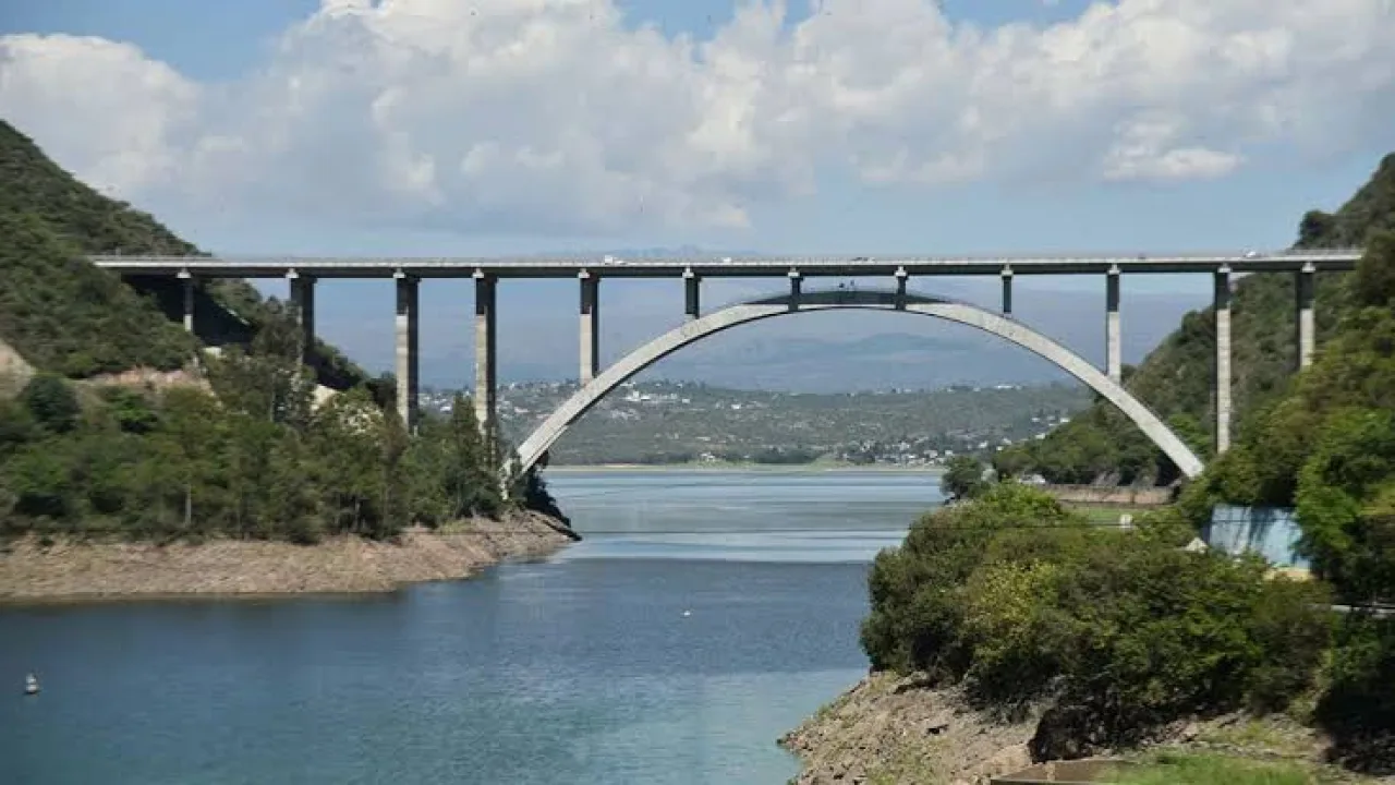 Puente de concreto con un arco central y carretera sobre un lago rodeado de vegetación y montañas en el fondo