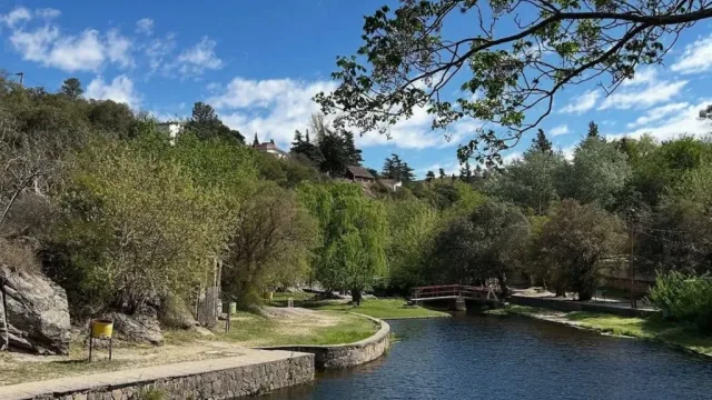 Río de corriente tranquila con murallas de piedra, un pequeño puente rojo, árboles y un cielo parcialmente nublado