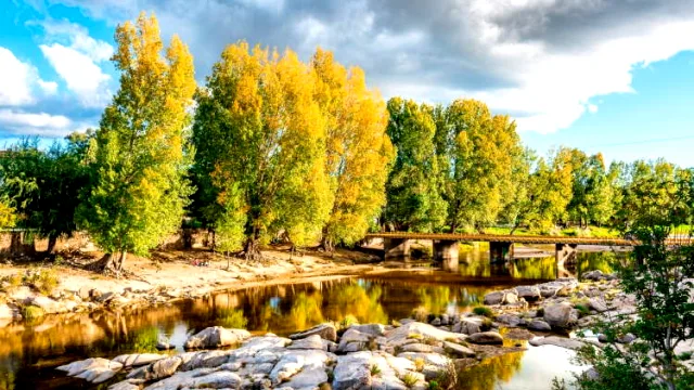 Curso de río con piedras en la orilla, puente pequeño y árboles con hojas amarillas y verdes reflejándose en el agua bajo un cielo parcialmente nublado
