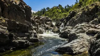 Río de aguas cristalinas corriendo entre grandes rocas y formaciones rocosas en las sierras con vegetación al fondo y cielo azul