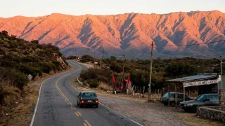Ruta asfaltada con un auto circulando, montañas y vegetación árida al fondo al amanecer
