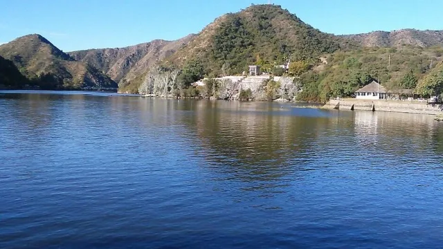 Panorámica de un lago con agua calma al frente y montañas cubiertas de vegetación en el fondo bajo un cielo despejado