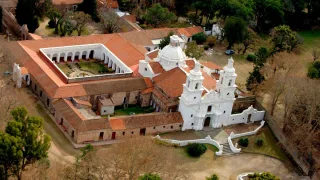 Edificio histórico con iglesia blanca de dos torres y patio cuadrado con galerías, rodeado de árboles y caminos de tierra