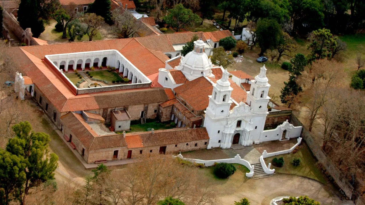 Edificio histórico con iglesia blanca de dos torres y patio cuadrado con galerías, rodeado de árboles y caminos de tierra