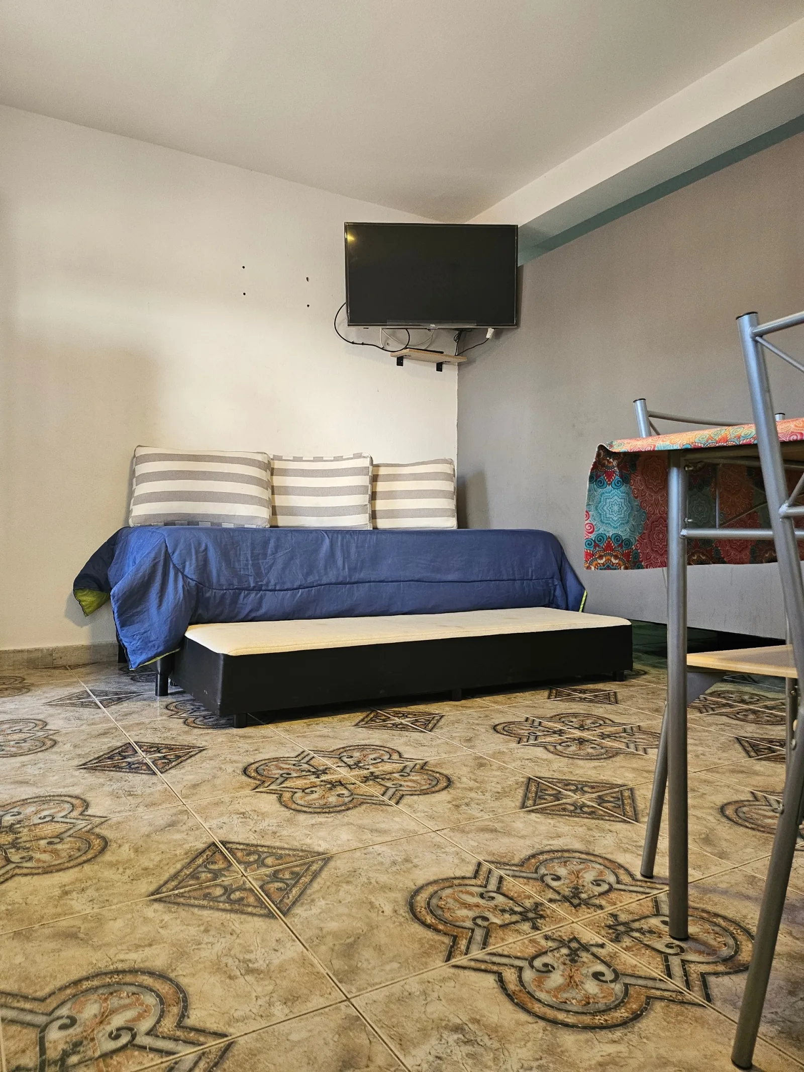 Sofá cama con tres almohadones a rayas, televisor colgado en la pared y mesa con mantel colorido y silla metálica