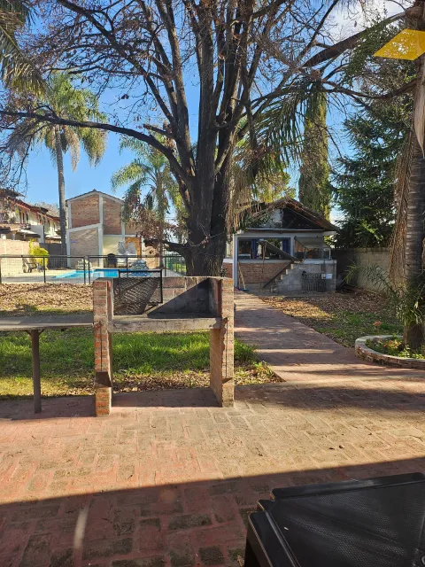 Vista de una cabaña al fondo con techo a dos aguas, frente a un jardín con parrilla y pileta cercada