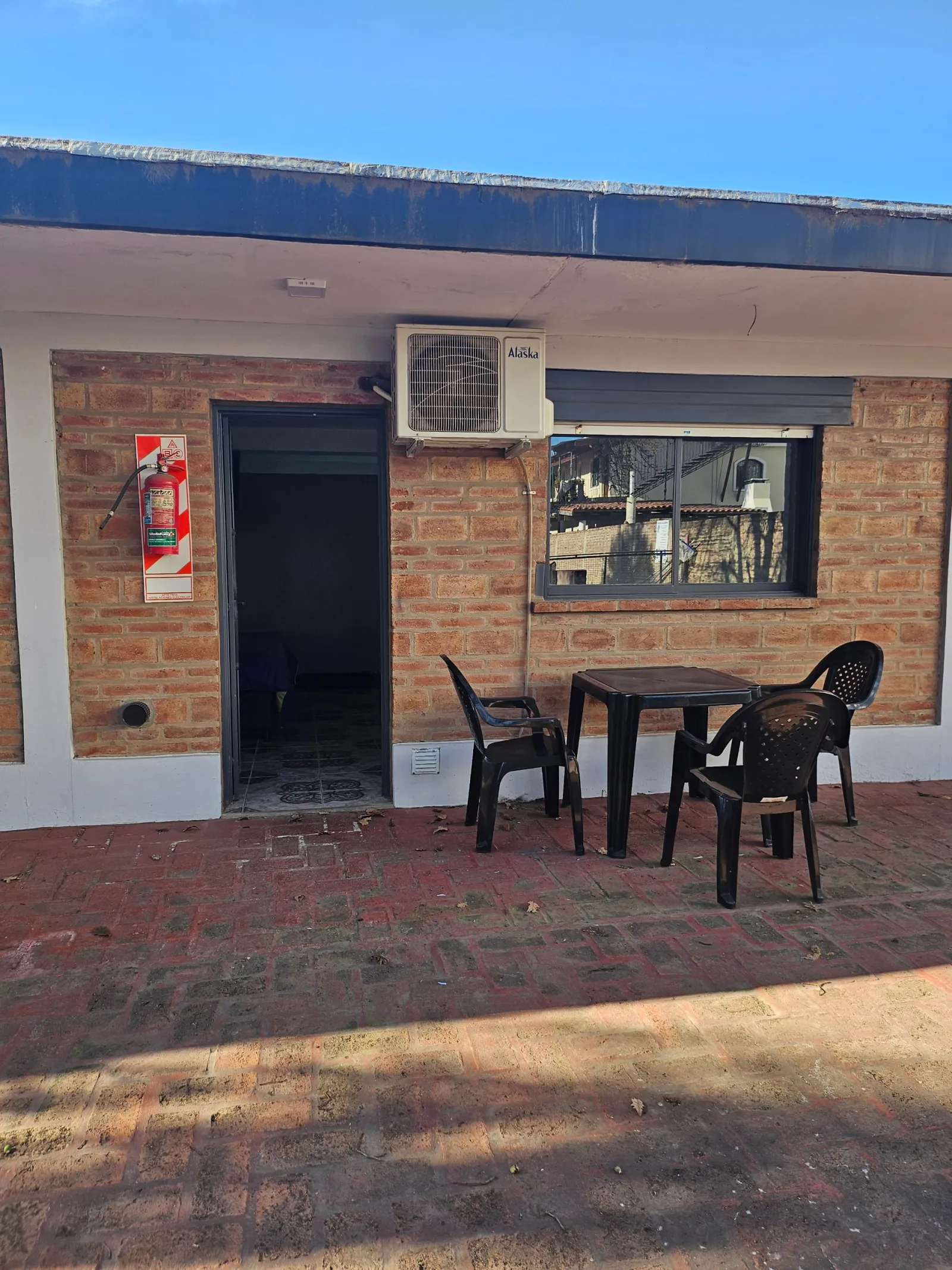 Patio con piso de ladrillo, una mesa negra con tres sillas de plástico y una entrada a cabaña con muros de ladrillo visto y una ventana con aire acondicionado