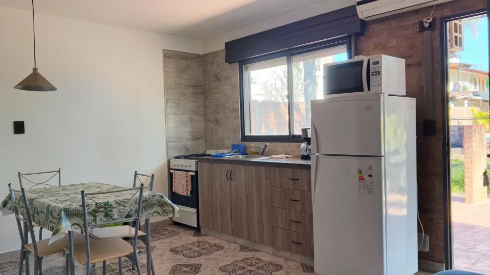 Cocina con alacena de madera, ventana grande, heladera blanca con microondas encima, mesa con mantel y cuatro sillas de metal, puerta vidriada que da al patio