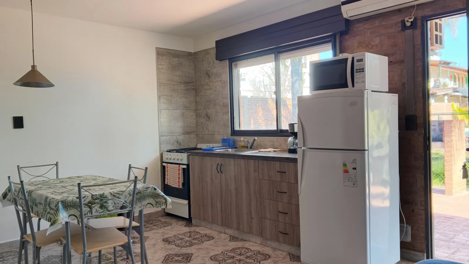 Cocina con alacena de madera, ventana grande, heladera blanca con microondas encima, mesa con mantel y cuatro sillas de metal, puerta vidriada que da al patio