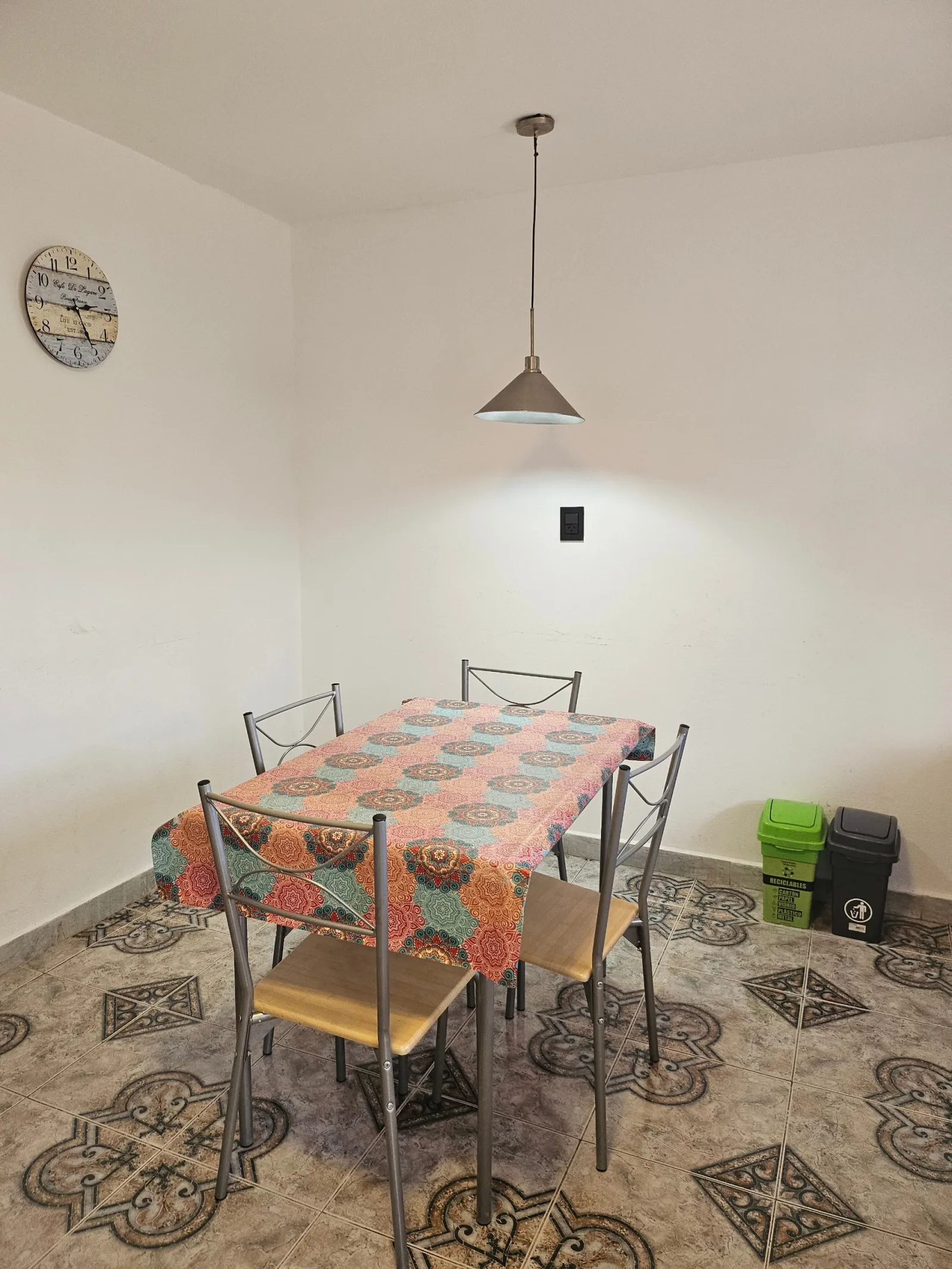 Mesa rectangular con mantel de colores y cuatro sillas metálicas, lámpara colgante y dos contenedores para separación de residuos