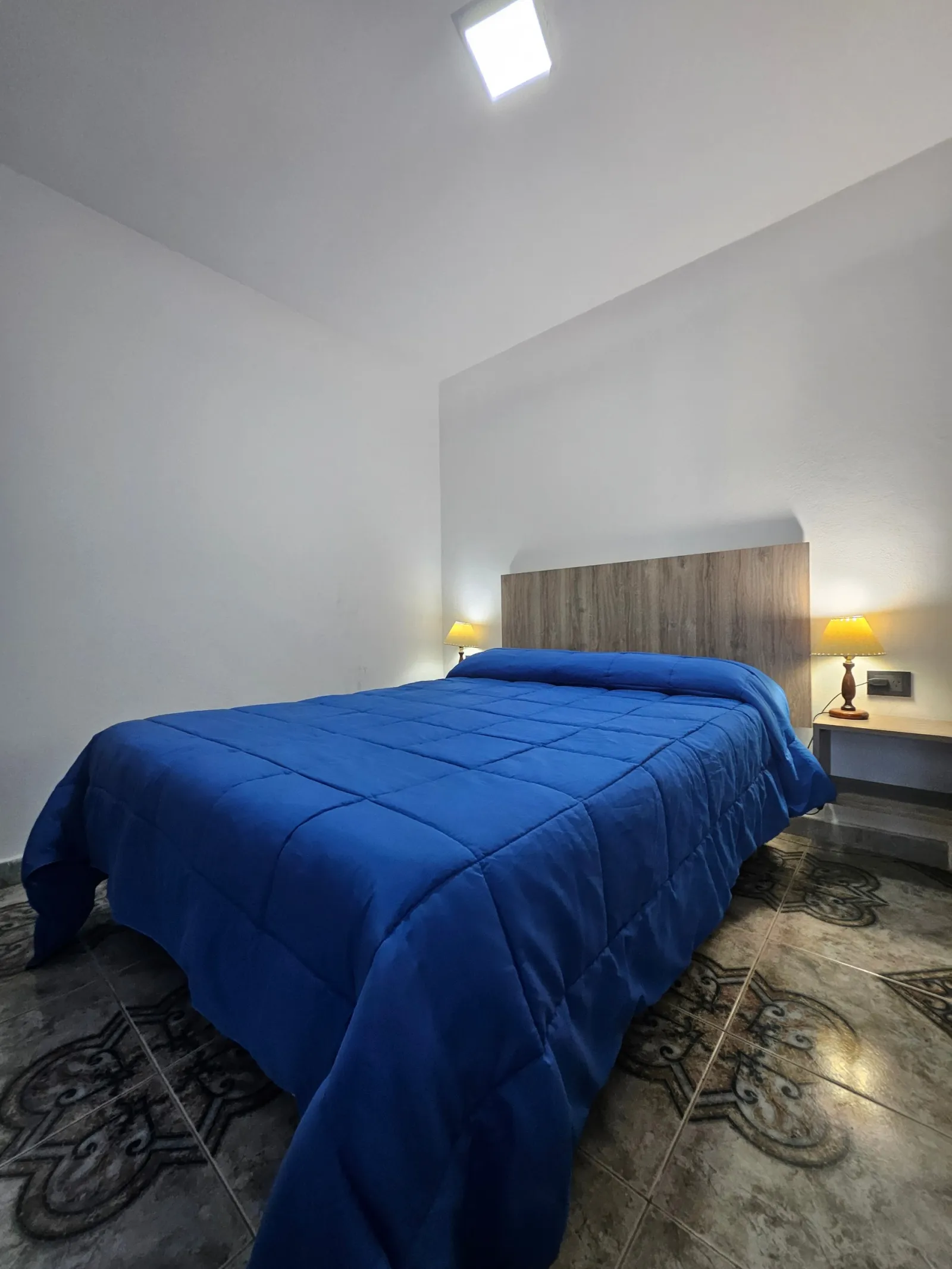 Habitación con cama matrimonial con cabecera de madera y acolchado azul, dos mesas de luz con lámparas encendidas