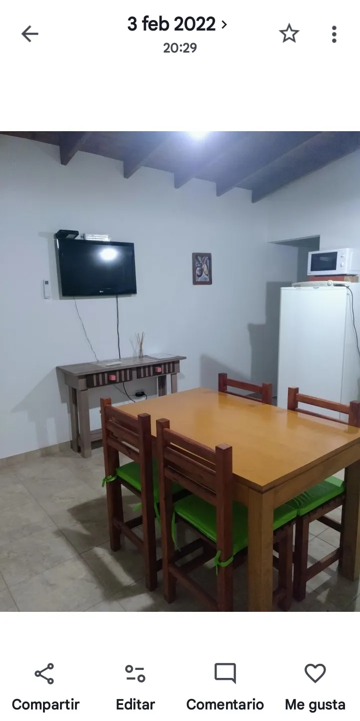 Mesa rectangular de madera con cuatro sillas, al fondo una mesa auxiliar, televisor colgado y heladera con microondas sobre ella