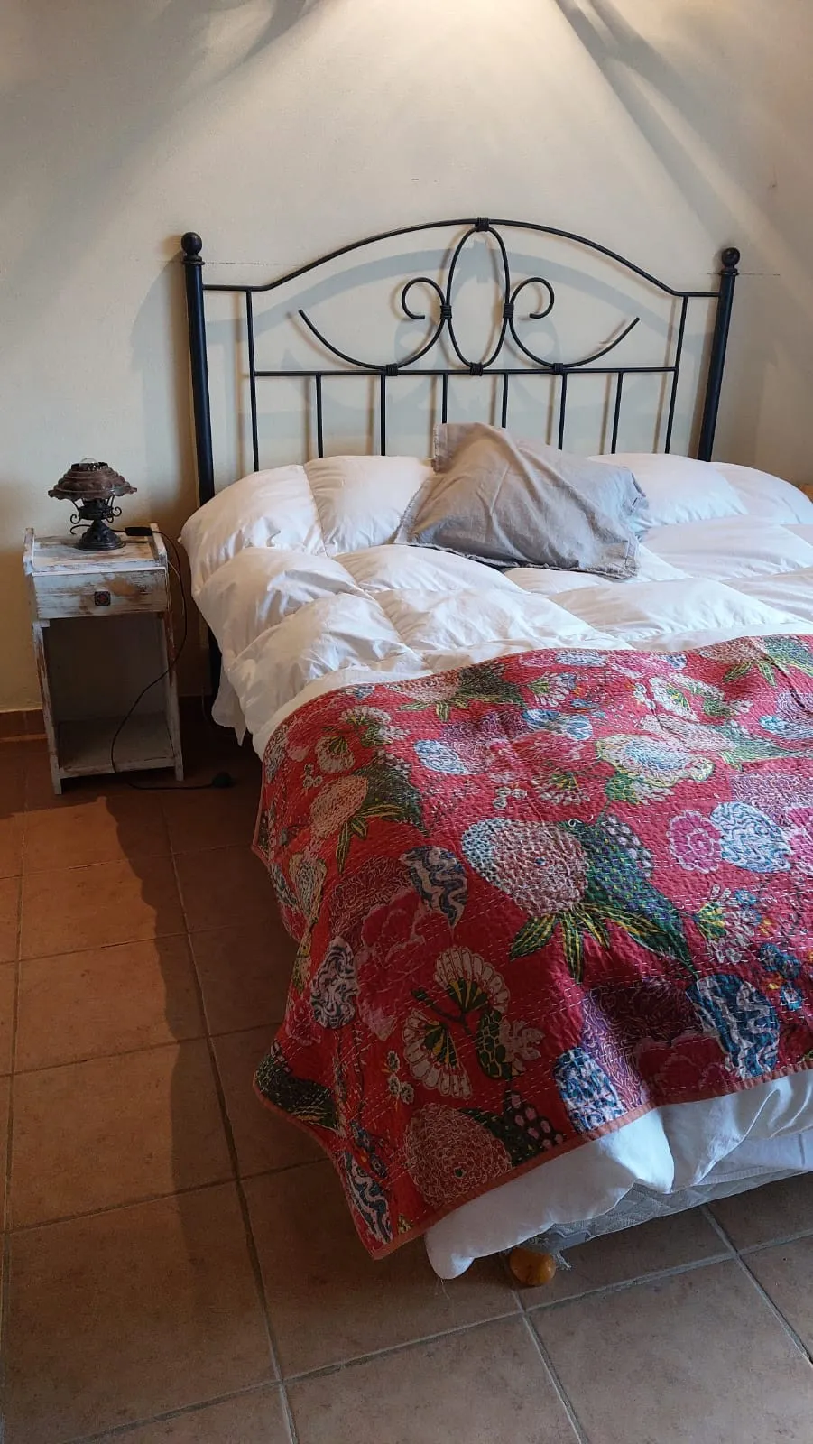 Cama con acolchado blanco, cobija estampada en tonos rojos y verdes, almohadón gris y mesa de noche de madera blanca desgastada