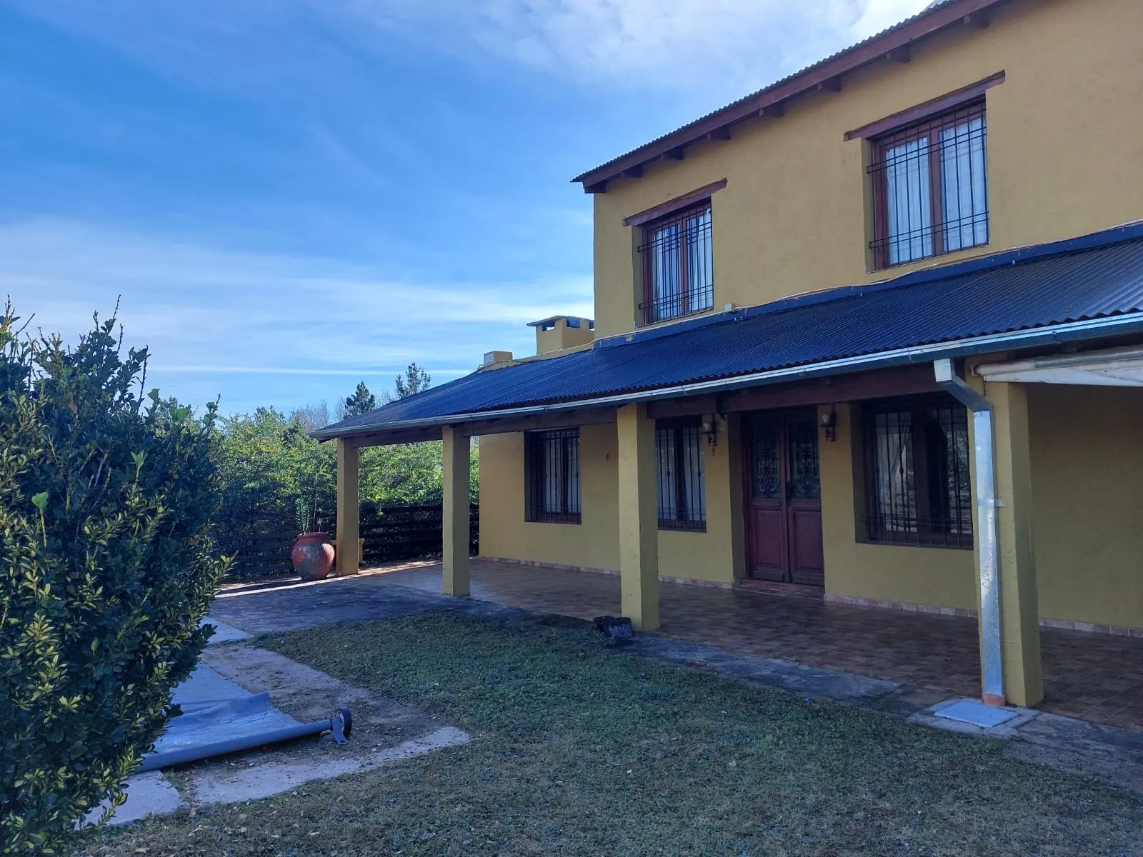 Casa de dos plantas con paredes amarillas, galería con techo de chapa, rejas en todas las ventanas y puerta de madera, jardín con césped y camino de piedra