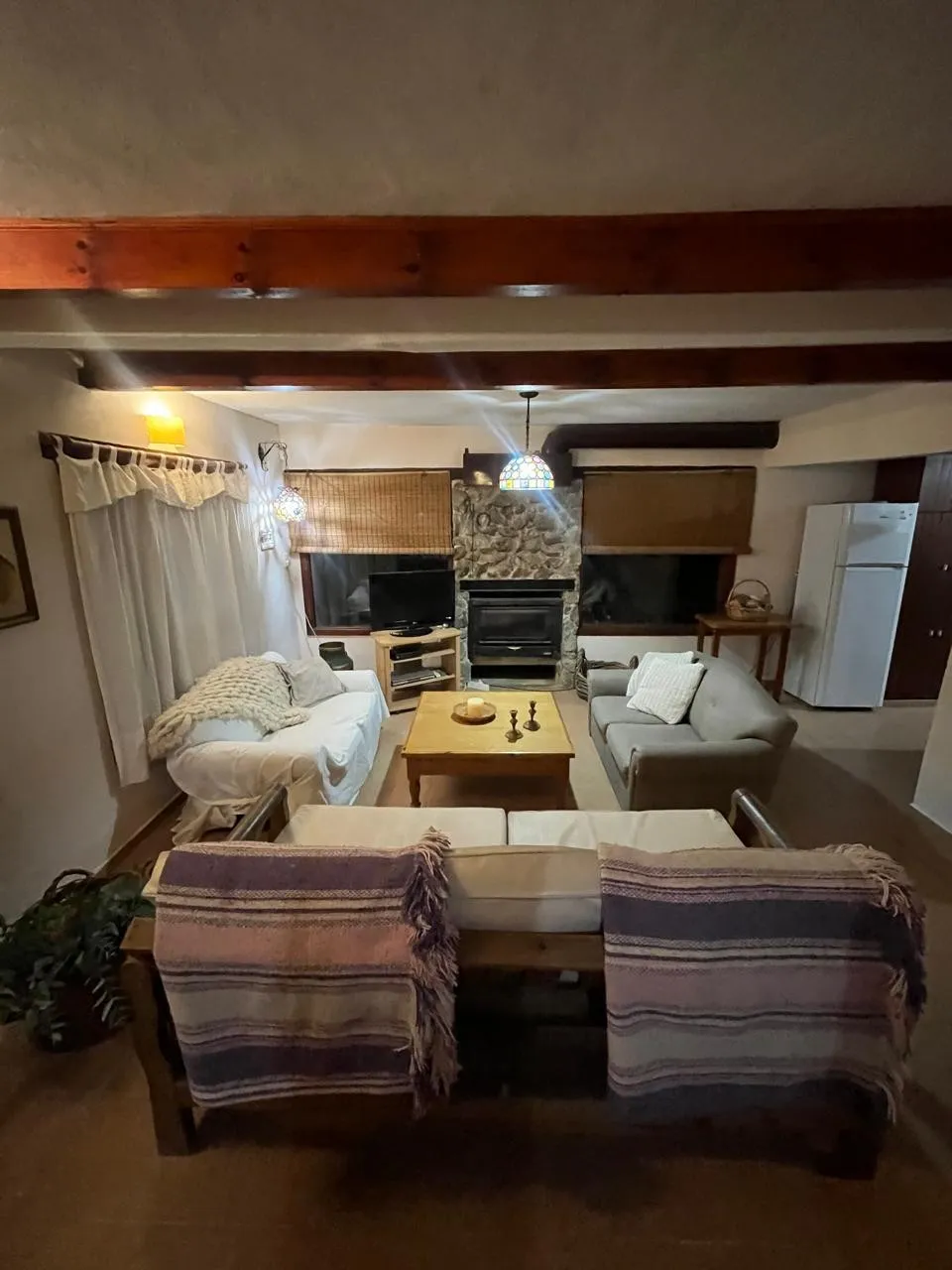 Living con tres sillones, mesa ratona de madera, hogar a leña revestido en piedra, dos ventanas con persianas y una cocina con heladera blanca al fondo