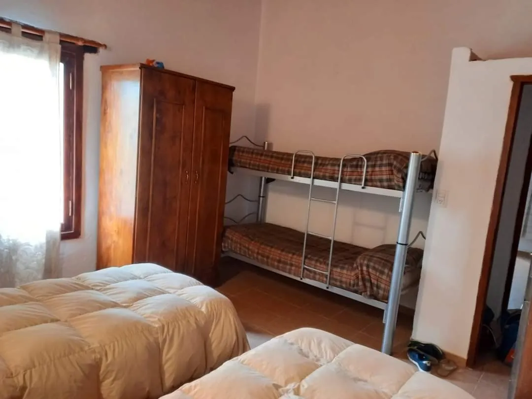 Habitación con una cama cucheta metálica, ropero de madera, dos camas con acolchados blancos y ventana con cortina blanca