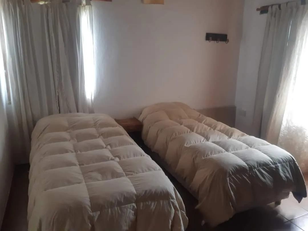Dos camas individuales con acolchados claros separadas por una mesa de noche en una habitación con paredes blancas y cortinas translúcidas.