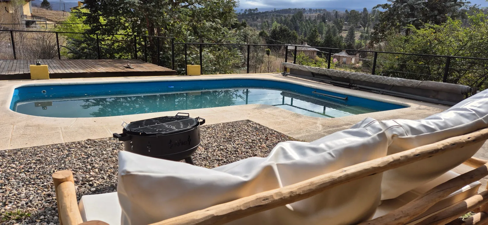 Pileta rectangular al aire libre rodeada por piedras y deck de madera, con un sillón con funda blanca y paisaje de árboles y cerros al fondo