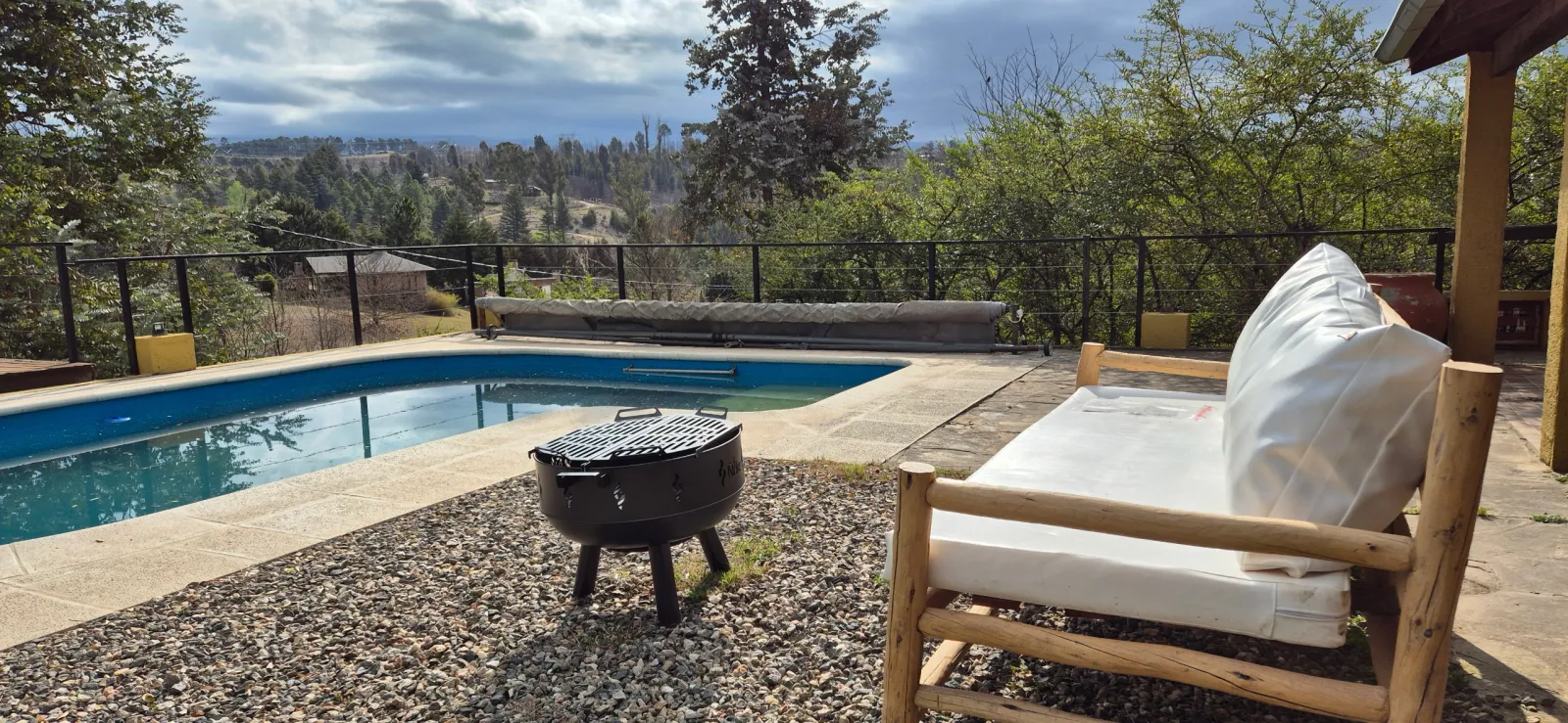 Pileta con borde de piedra, parrilla externa y sillón con cojines blancos en espacio exterior