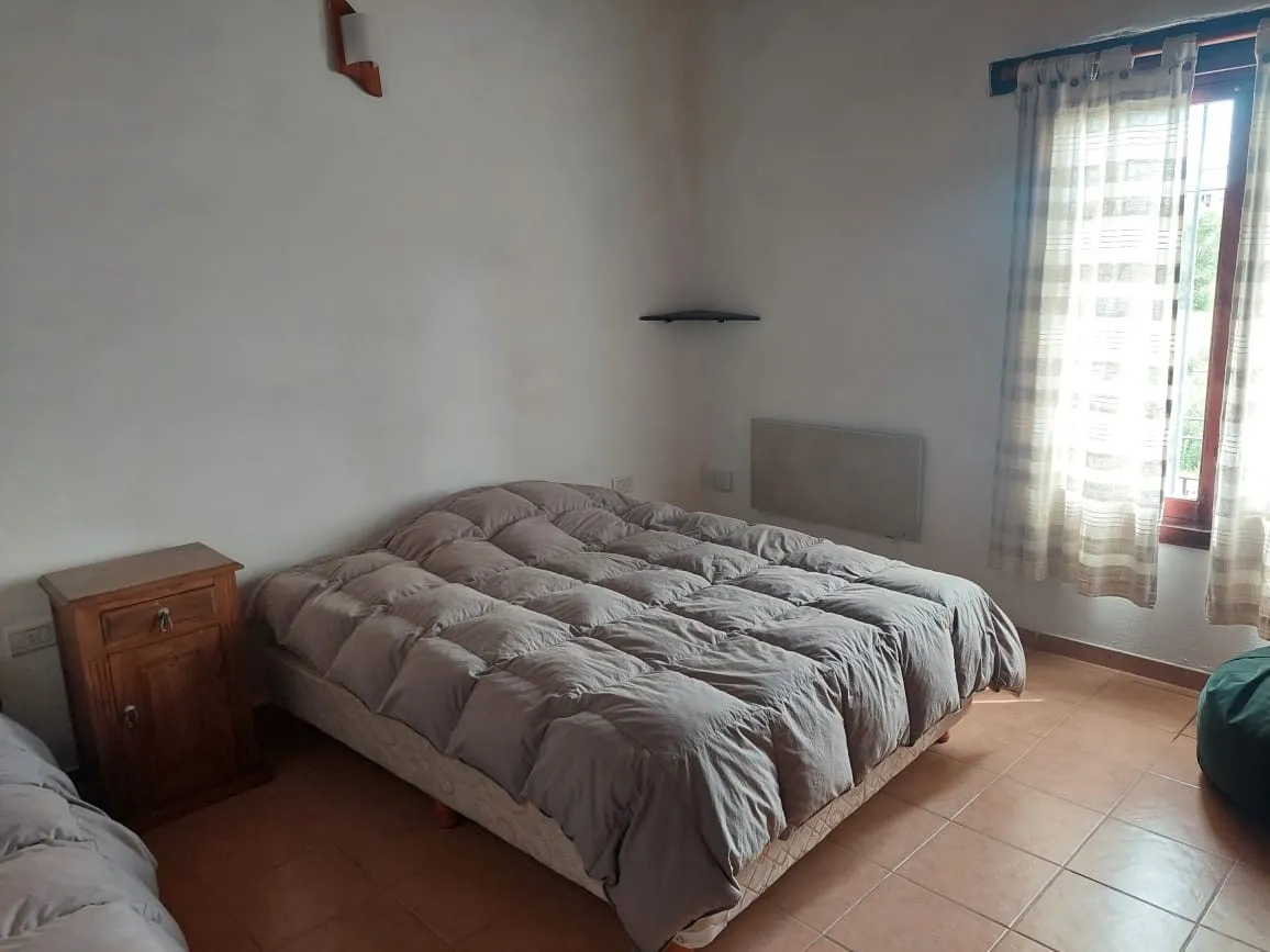 Habitación con cama doble con acolchado gris, mesa de luz de madera junto a la pared blanca, ventana con cortinas y luz natural entrante