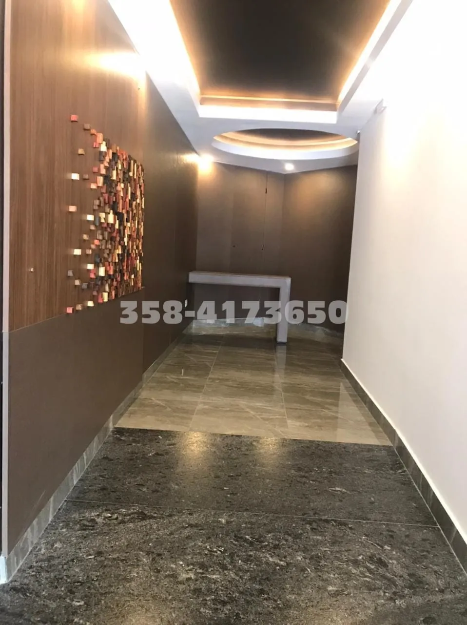 Pasillo con piso de baldosas en colores neutros, pared blanca a la derecha y pared marrón a la izquierda con cuadro artístico de piezas de madera y tres luces empotradas, consola blanca al fondo