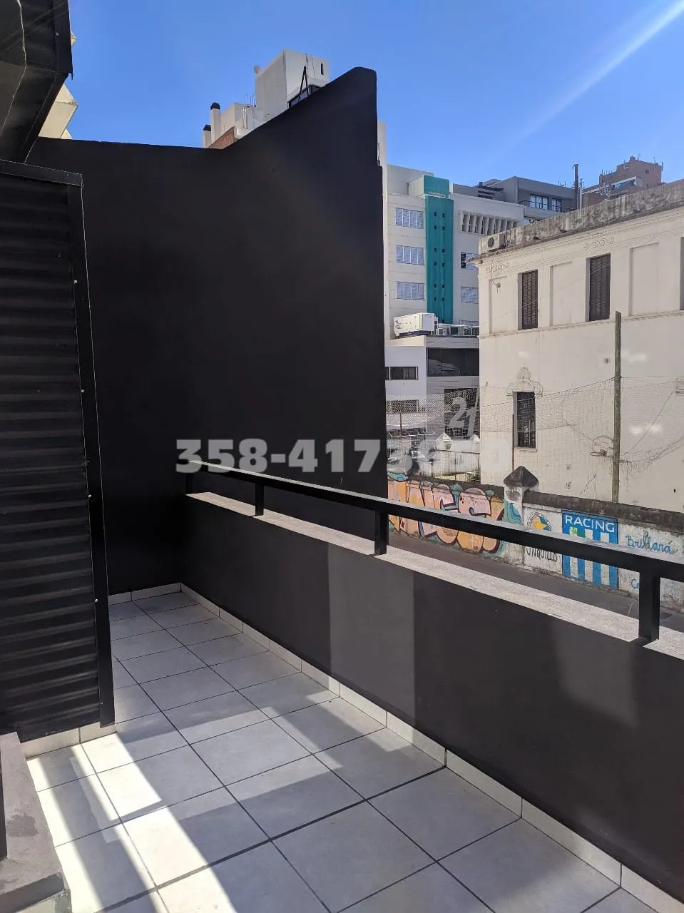 Balcón pequeño con baranda metálica negra, piso de baldosas claras y paredes oscuras, vista a edificios con grafitis en las paredes exteriores
