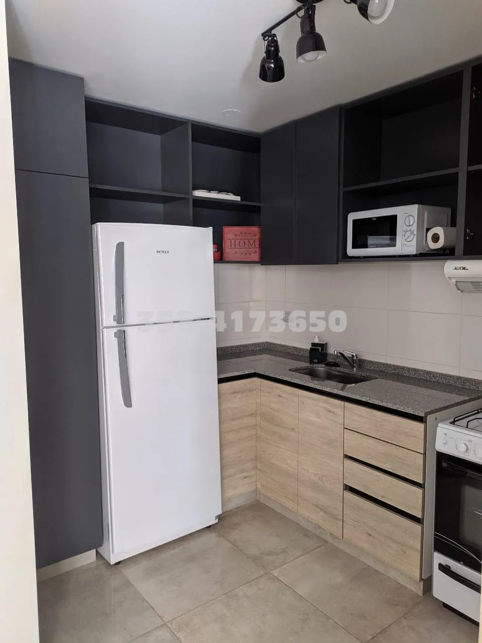 Cocina pequeña con heladera blanca, microondas sobre estante, mesada con bacha de acero inoxidable, muebles bajo mesada y alacenas negras