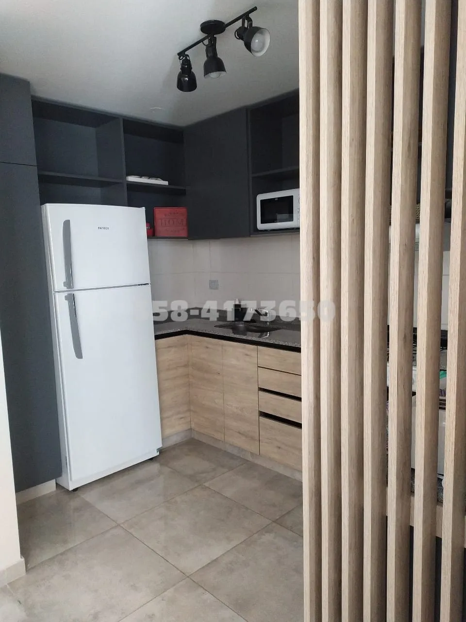 Cocina con mueble bajo de madera clara, mesada negra, heladera blanca, microondas blanco y estantes superiores en color oscuro, divisoria de listones verticales de madera