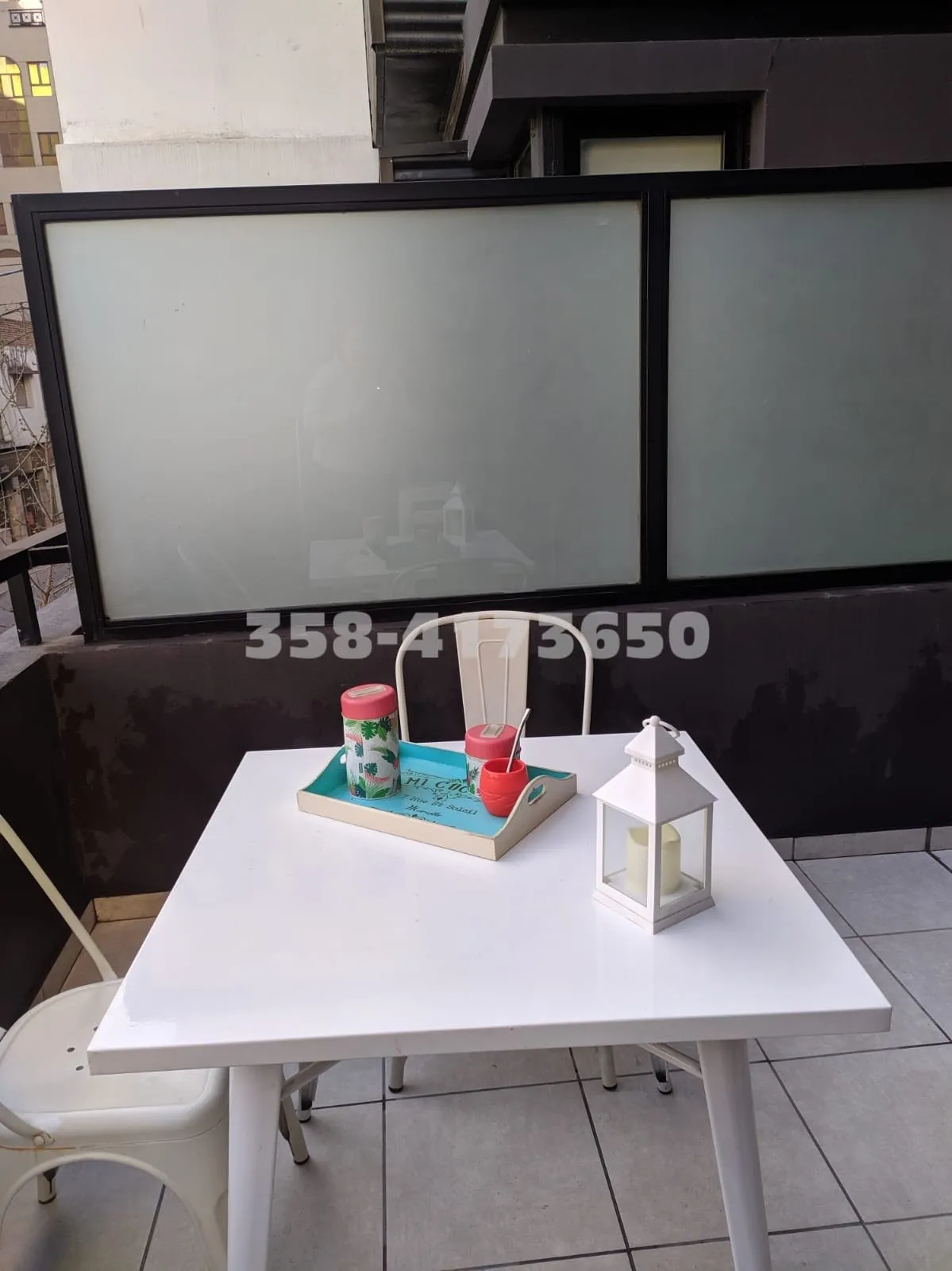 Balcón con mesa blanca, dos sillas metálicas blancas, bandeja con termo, mate y azucarera, y lámpara blanca con vela dentro