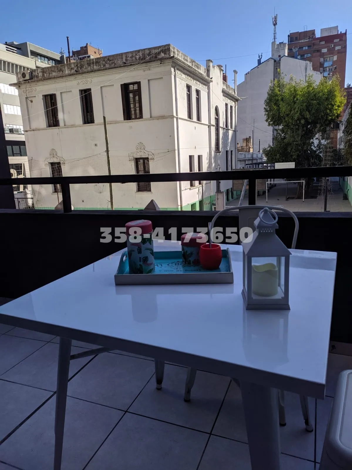 Mesa blanca cuadrada con bandeja que contiene termo, mate y azucarera, hay una linterna blanca decorativa al costado. Al fondo se ven edificios y árboles.