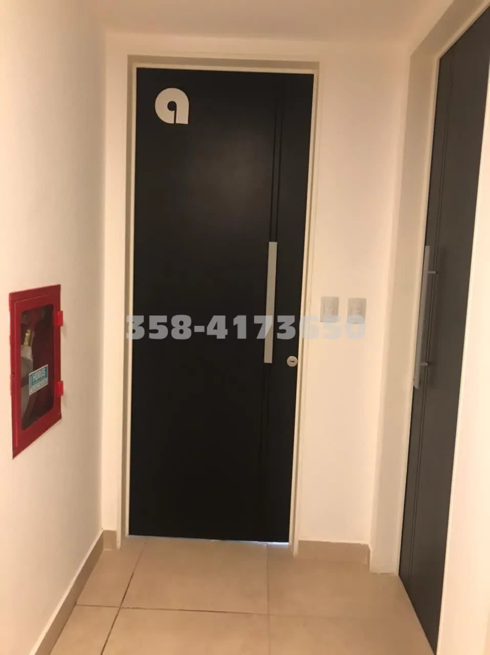 Puerta negra con manija plateada en pasillo de paredes blancas y detalle de caja roja a la izquierda