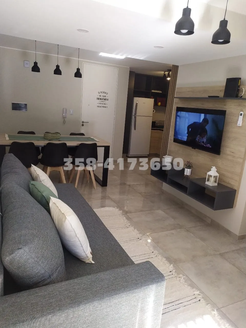 Living comedor con sofá gris, mesa con sillas negras, cocina al fondo y televisor colgado en la pared