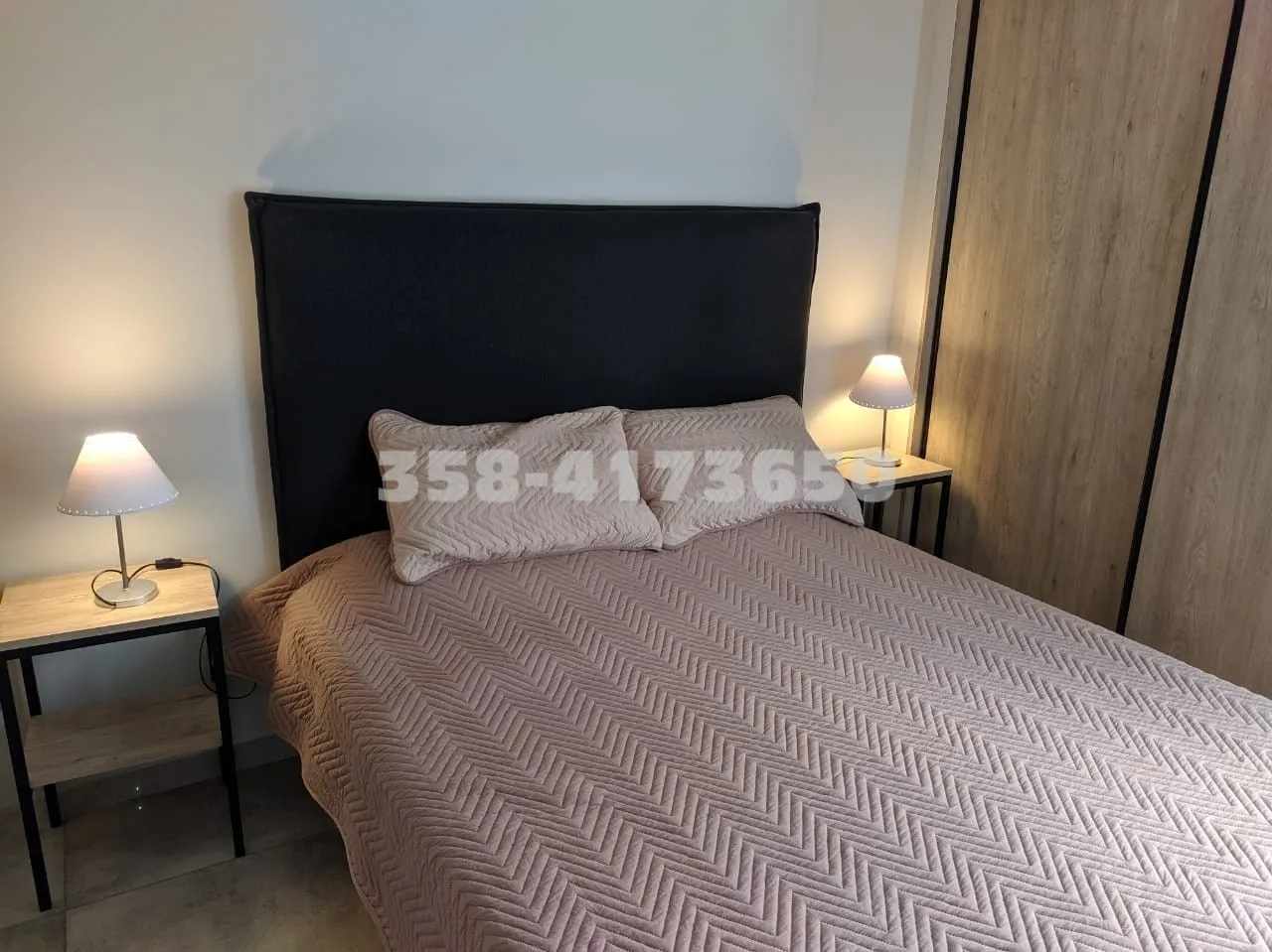 Cama matrimonial con cabecera oscura, dos almohadas con funda y cubrecama beige, mesas de luz de madera y lámparas encendidas a ambos lados