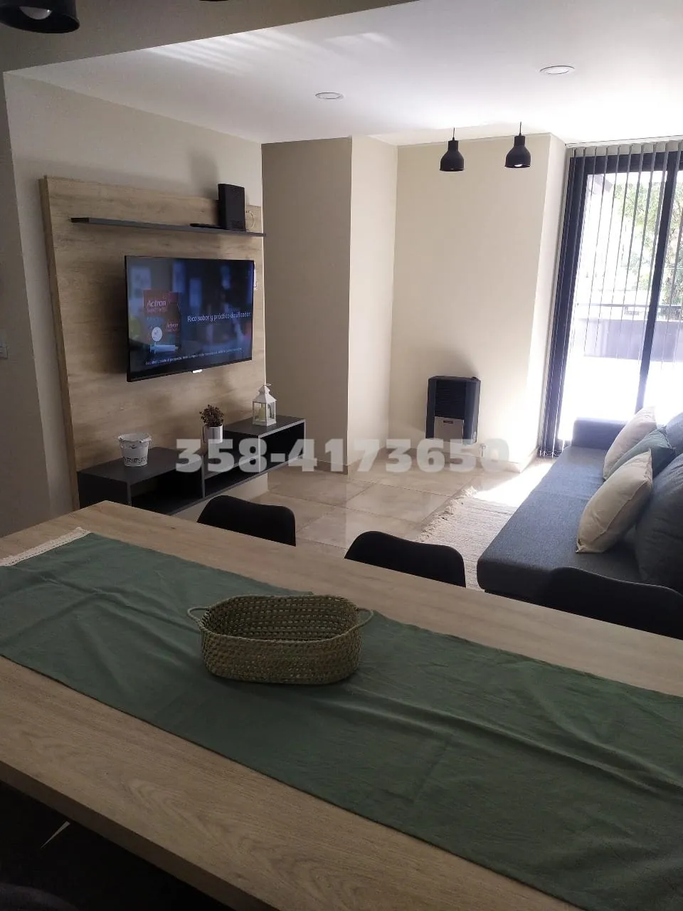 Sala de estar con sofá gris, televisor colgado en la pared y vista parcial a un balcón con cortina de vidrio