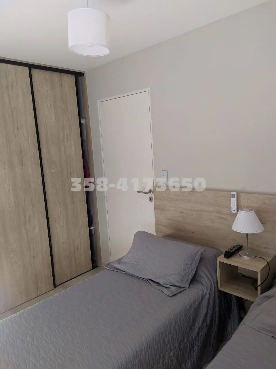 Habitación con cama con acolchado gris, lámpara de mesa, control remoto y placard de madera clara con puerta semi abierta
