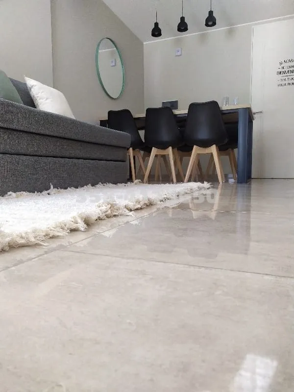 Piso de baldosas beige claro con alfombra blanca de flecos, sillón gris a la izquierda y sillas negras con patas de madera