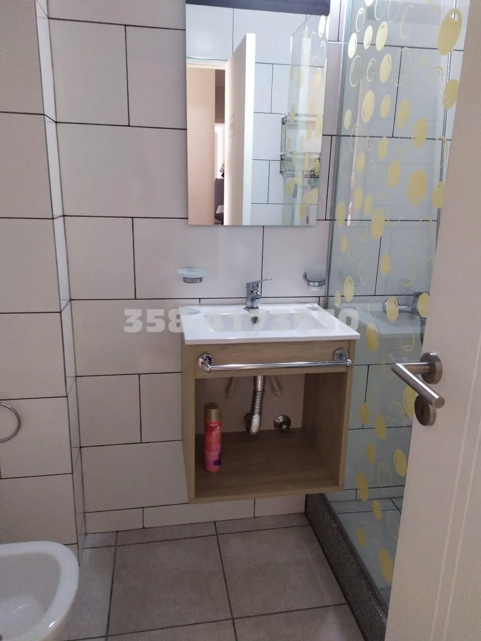 Baño con espejo rectangular y mueble bajo con lavabo blanco, paredes revestidas con azulejos beige y ducha con mampara de vidrio con puntos amarillos