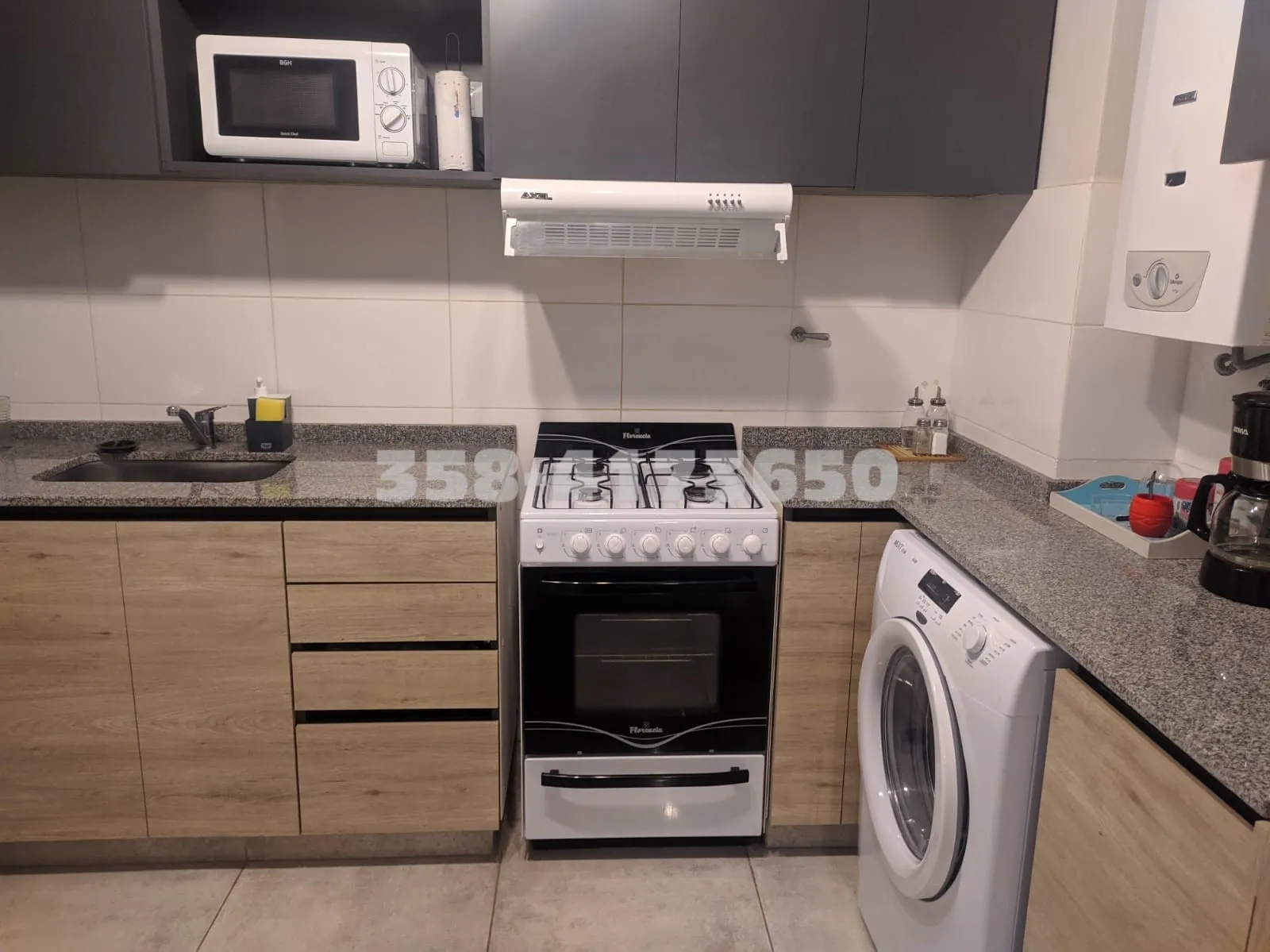 Cocina equipada con muebles bajos de madera clara, horno con anafe a gas, campana extractora, microondas, lavadora blanca y mesadas de granito gris
