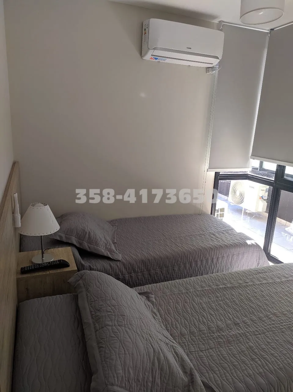 Habitación con dos camas individuales con acolchados grises, lámpara blanca sobre mesa de luz y ventana con cortinas enrollables que permite entrada de luz