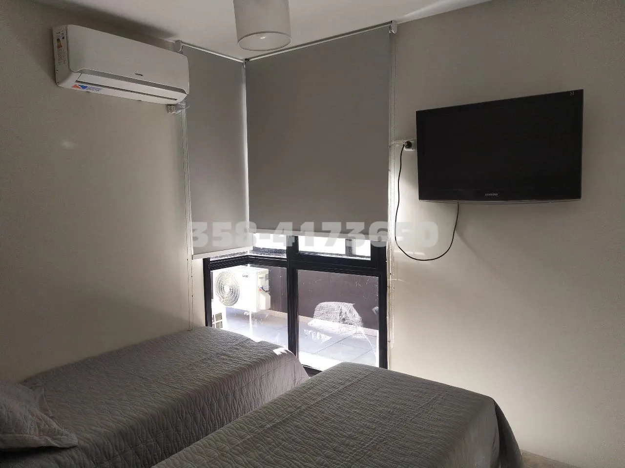 Habitación de un departamento con dos camas individuales cubiertas con colchas, ventana con cortina, acondicionado instalado en la parte superior y TV colgada en la pared frente a las camas.