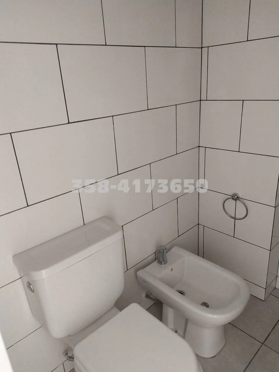 Baño pequeño con paredes cubiertas de azulejos blancos, inodoro blanco y portarrollos de toallas de mano metálico